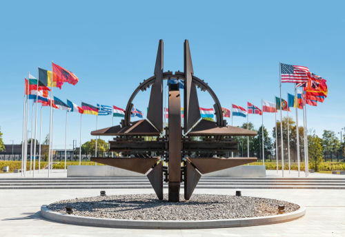 shutterstock_1399317887.jpg NATO emblem with flags of member countries in background.
