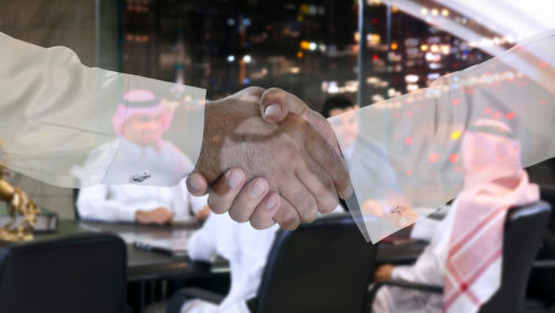 shutterstock_1174896658 Two people shaking hands; men in background discussing.