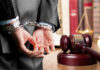Person in handcuffs near gavel and scales of justice.