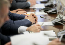 AI Spending Sucker-Punches IBM Business meeting participants with hands clasped at a conference table with microphones