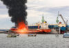 A large ship on fire with black smoke rising, surrounded by smaller fishing boats in a harbor