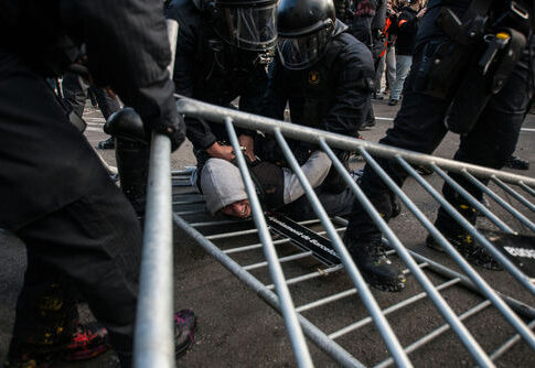Police in riot gear detain a person on the ground near a toppled metal barricade