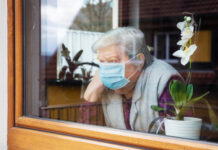 Elderly woman wearing mask, looking out window beside orchid.